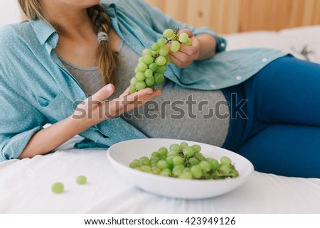 Young pregnant woman holding green grapes with a plate standing beside her. Pregnant woman eats healthy vegan food.