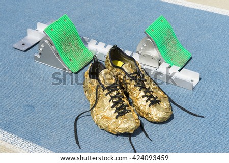 Gold running shoes sit next to sprinting starting blocks on a textured blue and tan running track