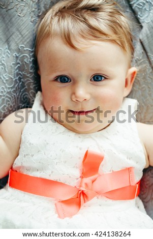 Portrait of cute adorable Caucasian smiling laughing baby girl with blue eyes  in white dress with red bow lying on bed looking in camera, shot from above, overhead, top view