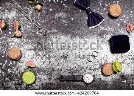 Wedding day concept. White gold luxury necklace, macaroones, two wedding rings on the velvet box, deep blue bowtie, roses, lipstick and men's watch on the table. Place for text.
