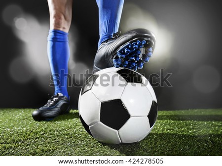 close up legs and feet of football player in blue socks and black shoes playing  with the ball standing on green grass pitch isolated on black background with flash lights and lens flare