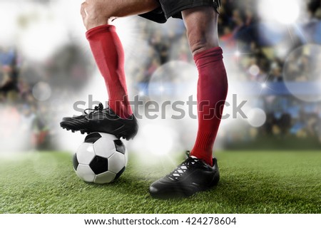 close up legs and feet of football player in red socks and black shoes playing with the ball standing on stadium green grass pitch with background flashes and light flares