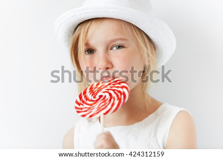 Close up image of happy little girl holding big spiral lollipop. Adorable blonde preschool child wearing fashionable white hat and dress, looking away, licking candy. Selective focus, film effect