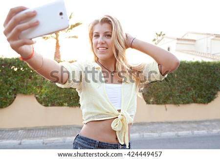Portrait of a beautiful blond teenager using a smart phone to network, taking selfies pictures of herself in a suburban home exterior at sunset, outdoors. Technology lifestyle, adolescent flirting.