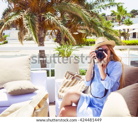 Happy young blond woman taking photo with camera on outdoor patio sofa furniture. Beautiful woman at hotel resort terrace smiling for picture enjoying modern luxury living