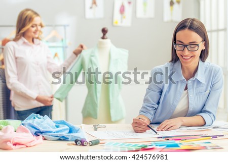 Beautiful designer is working in dressmaking studio, drawing sketches and smiling, in the background another designer is measuring jacket
