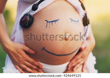 Pregnant woman belly closeup with smiling funny face drawing listening to the music outdoors. Earphones at woman's belly. Happy pregnancy, fun, creativity. Art, Music therapy. Smile face at belly. 