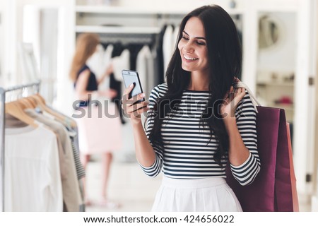Technologies make shopping easier. Beautiful young woman with shopping bags using her smart phone with smile while standing at the clothing store  Royalty-Free Stock Photo #424656022