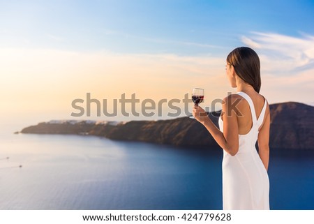 Young woman drinking red wine on outdoor terrace watching beautiful sunset view of Mediterranean Sea. Female in white sundress on summer Europe travel vacation in Santorini, Greece.