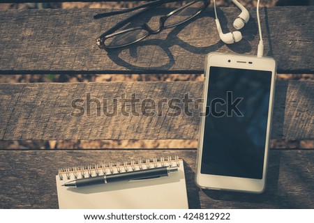 Smartphone with blank area on touchscreen with in ear headset, notebook, pen, glasses on rustic wood table in morning time with vintage filter effect. Modern outdoor lifestyle with technology concept.