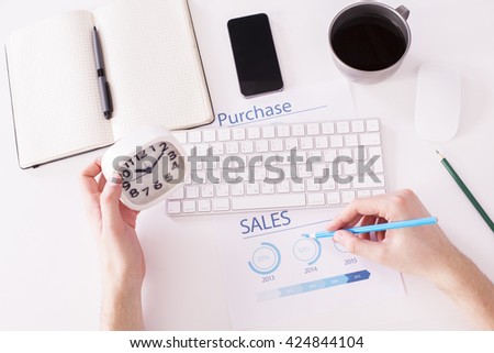 Top view of white office desktop with male hands holding clock and drawing pie-chart on  business report paper