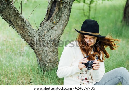   Wearing a white sweater and black hat in blooming garden. Beautiful young woman with long wavy hair thin slender figure perfect body and pretty face natural make-up. Photographer making pictures.