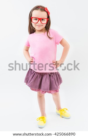 Cute fashion-monger girl in pink dress tshirt and skirt red glasses frames. Trendy kid with empty no image tshirt you can place your own text information or ad logo. Studio shot. White background