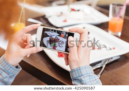 Hands of young woman using mobile phone and taking pictures of chocolate dessert on white plate in cafe