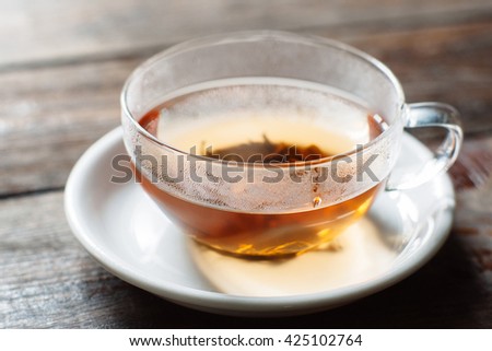 Cup of hot black tea close-up. Glass cup on saucer front view. Cup with tea leaves on dark wooden background. 