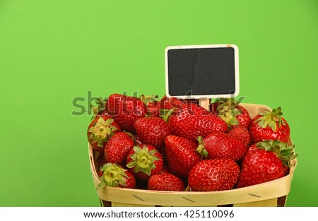 Mellow fresh red summer strawberries in wooden wicker basket with chalk blackboard price tag sign over green paper background, high angle view