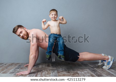 Smiling bearded father doing push-ups with little son sitting on his back over grey background Royalty-Free Stock Photo #425113279