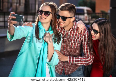 two young women and the guy taking selfie with mobile phone. The concept of a Merry mood and laughter on the street. Outdoor lifestyle portrait