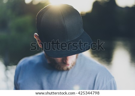 Photo Bearded Young Man Wearing Black Blank Cap. Green City Park Lake Background and Sunset effect. Front view. Horizontal Mockup. Royalty-Free Stock Photo #425344798