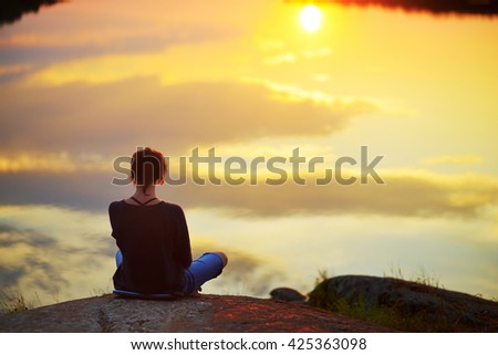 Young woman sitting on the stone enjoying peaceful moment of sunset. In the reflection of the lake water sees clouds and sun. Inner mind open, harmony life up in dusk sky, destiny, god, zen concept Royalty-Free Stock Photo #425363098