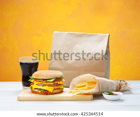 Fast food. Brown wrapping paper package with copyspace. Hamburger, potato fries, cola drink. Takeaway food. Wrapped French fries, packaging, Cola glass, tomato sauce, double cheese hamburger at wood.