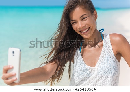 Cheerful young woman taking fun mobile phone selfie photos of herself on beach holidays during summer tropical travel vacation. Happy healthy Asian mixed race beauty girl smiling at smartphone.