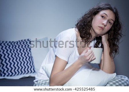 Sleepless young woman in pyjamas sitting on a bed with a white mug in her hand, with a tired look on her face Royalty-Free Stock Photo #425525485