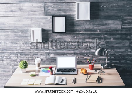 Modern designer workplace with blank white laptop, various stationery items, food and three blank picture frames hanging above on wooden wall. Mock up