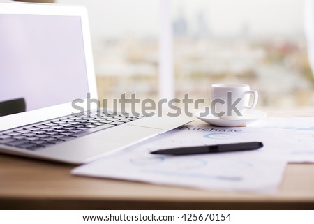 Closeup of office desktop with blank laptop screen, business report, coffee cup and pen on blurry city background. Mock up