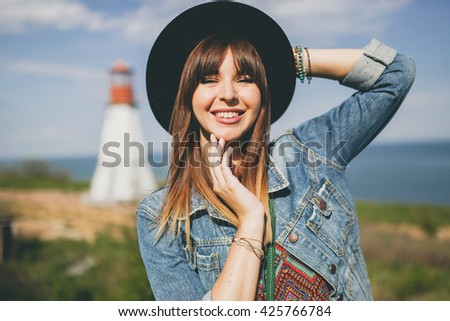 young woman sitting in field, nature background, lighthouse, hipster style, bohemian outfit, denim jacket, black hat, smiling, happy, summer, sunny, stylish accessories, bracelets, hands