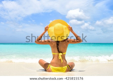 Happy carefree woman relaxing sitting in sand enjoying tropical beach destination. Perfect paradise summer vacation happiness. Back view of bikini girl holding yellow fashion hat on Caribbean holiday. Royalty-Free Stock Photo #425815576