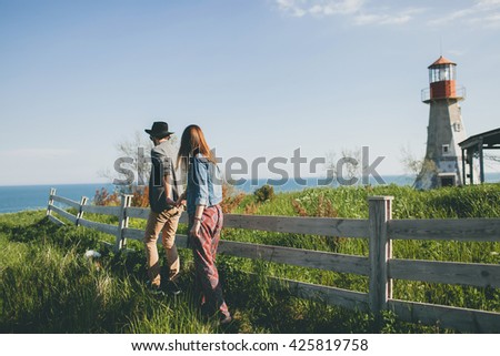 young couple hipster indie style in love walking in countryside, holding hands, lighthouse on background, warm summer day, sunny, bohemian outfit, hat