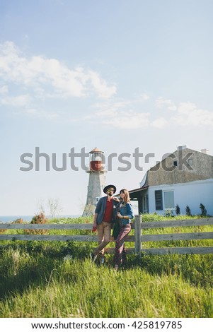 young couple hipster indie style in love walking in countryside, holding hands, lighthouse on background, warm summer day, sunny, bohemian outfit, hat