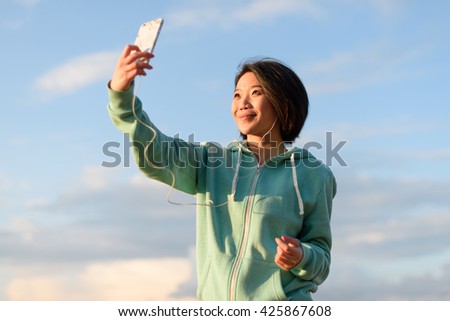 Pretty japanese woman taking selfie outdoor using her phone. Blue cloudy sky background