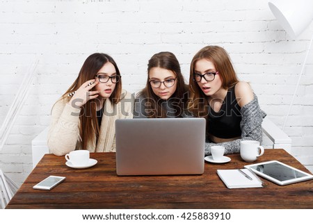 Three girls have coffee with laptop. Young girls friends watch something in internet. Shocking news. Girls are anxious, scared, surprised. Women and computer,  internet news, gossip concept. 