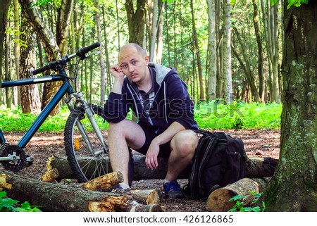 Cyclist with backpack, young man thinking with fatigue on face, in beautiful forest, summertime journey