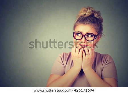 Closeup portrait nervous stressed young nerdy woman in glasses biting fingernails looking anxiously craving something isolated gray background. Human emotion face expression feeling reaction 