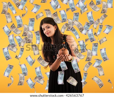 Pretty Indian/Asian young girl catching falling indian currency with terracotta piggy bank and smiling, standing isolated over yellow background