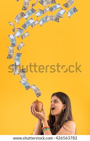 Pretty Indian/Asian young girl catching falling indian currency with terracotta piggy bank and smiling, standing isolated over yellow background