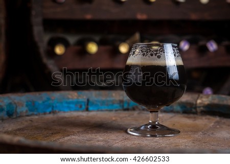 Snifter glass full of dark ale standing on a wooden barrel in a cellar Royalty-Free Stock Photo #426602533