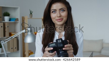 Smiling brunette woman wearing white shirt holding camera and looking at you, office interior at background.