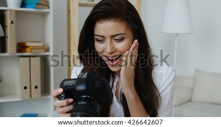 Cute young smiling brunette wearing white shirt sitting holding camera, office interior at background.