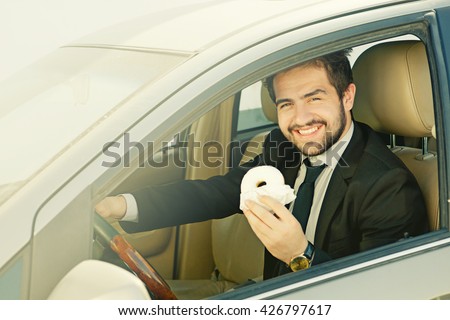 Toned picture of happy businessman in black business suit eating donut while driving car to his office. Freelance man smiling for camera.