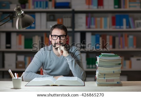 Confident adult student at the library late at night, he is sitting at desk and reading a book with hand on chin, learning and education concept