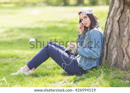 Teenage girl sitting in a park leaning on a tree and listening to music on headphones