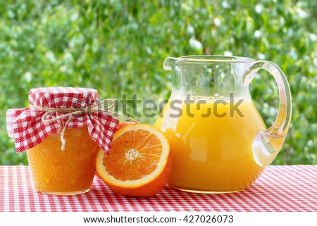 Orange, juice and jam  on a red checkered tablecloth. Background of green trees. 