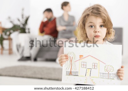 Upset little boy holding a drawing of a house, with his parents sitting angry on a couch in the blurry background