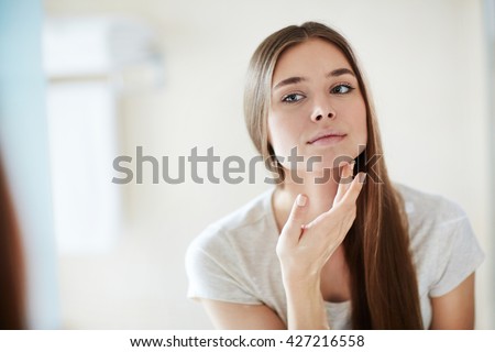 Young woman looking at mirror at home and applying cream on her face Royalty-Free Stock Photo #427216558