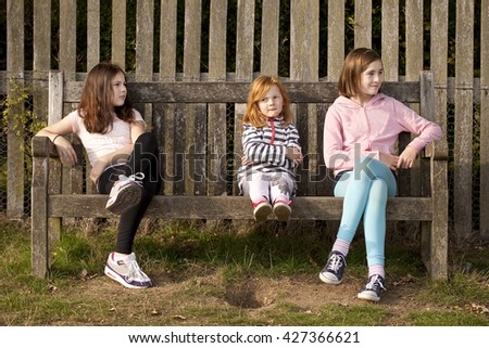 Three Sisters. Three sisters sit on a bench in a park. The elder sisters are looking at something in the distance but the youngest sister is bored and wants to get moving again. 
