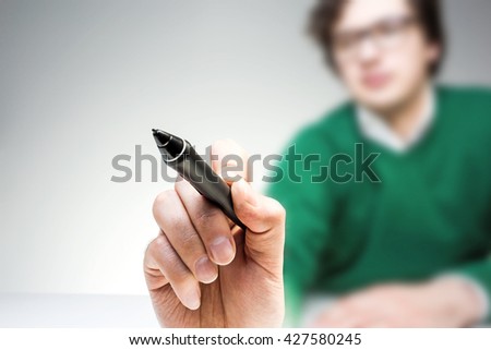 Blurry man in green pullover at white desk writing something with marker on light background